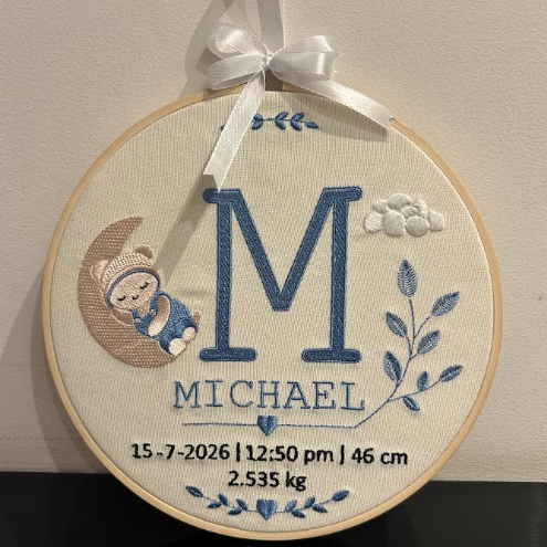 A personalized circular embroidery hoop ornament hanging on a wall, featuring a large blue embroidered letter M, the name MICHAEL, birth date and weight, a crescent moon with a baby figure, and blue floral details, tied with a white ribbon.
