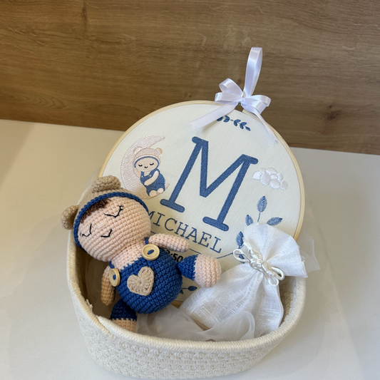 A personalized baby keepsake gift set featuring a crocheted doll dressed in blue and brown, nestled inside a round white embroidered hoop with a custom monogram 'M' and the name 'MICHAEL', complete with a white drawstring bag and hanging ribbon.
