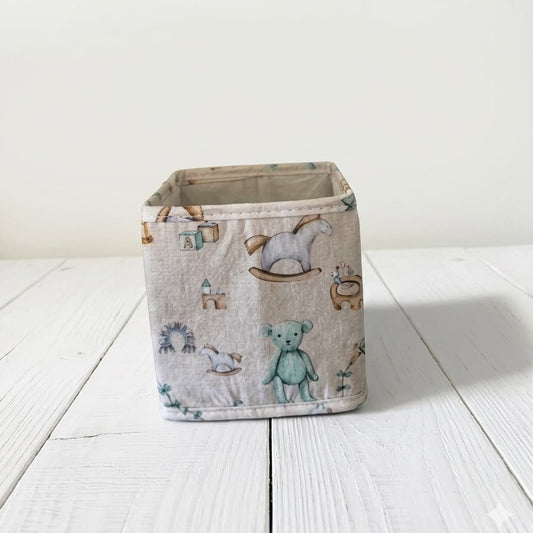 A square fabric storage basket with a light beige background and a whimsical print of teddy bears, rocking horses, and small houses in muted blue, green, and brown tones, set on a white wooden surface against a white wall.